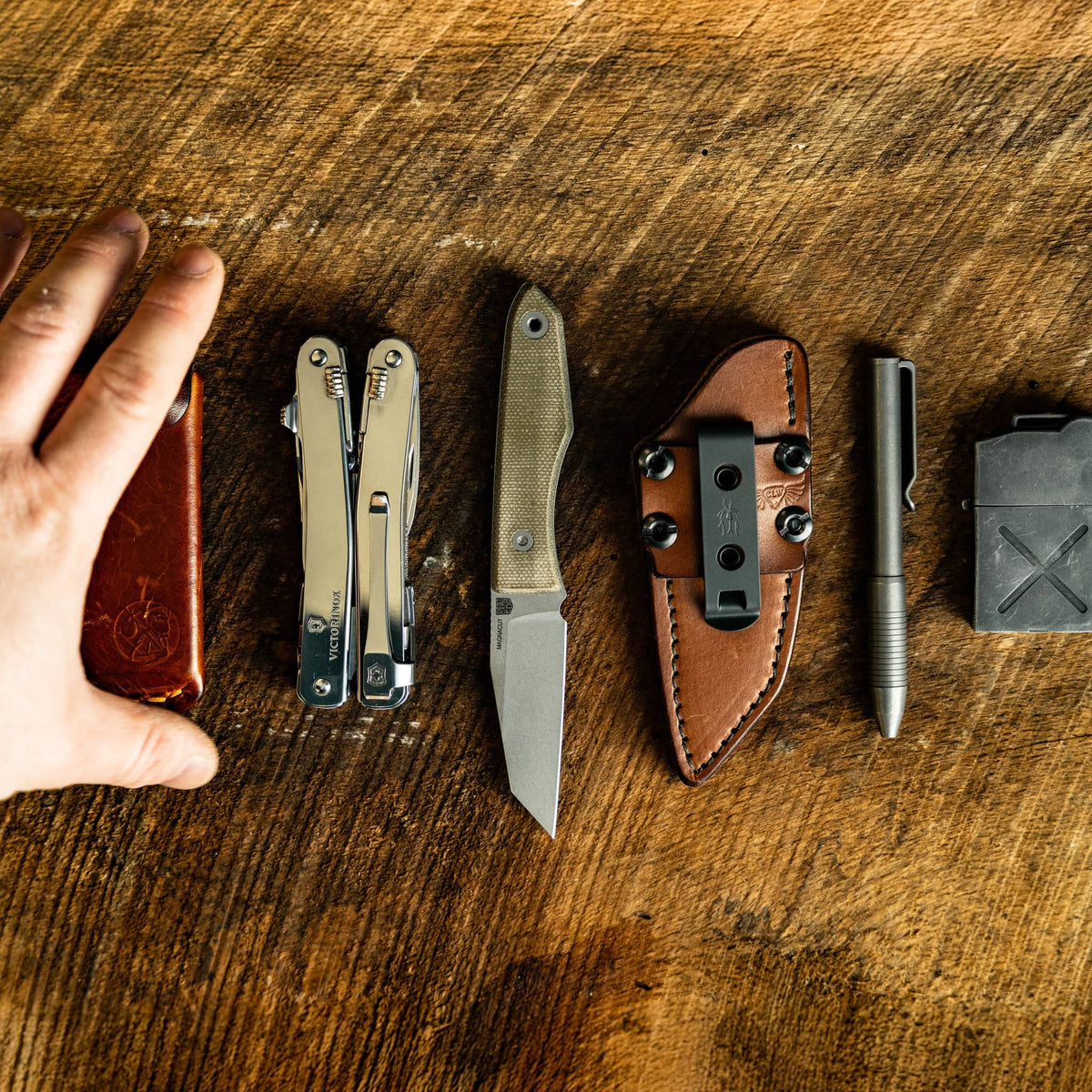Hand and gear: Lookout knife and leather sheath arranged beside everyday carry gear, with a hand for scale