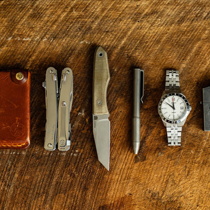 EDC lineup: Lookout reverse tanto knife beside a multitool, pen, and wristwatch on a wooden surface