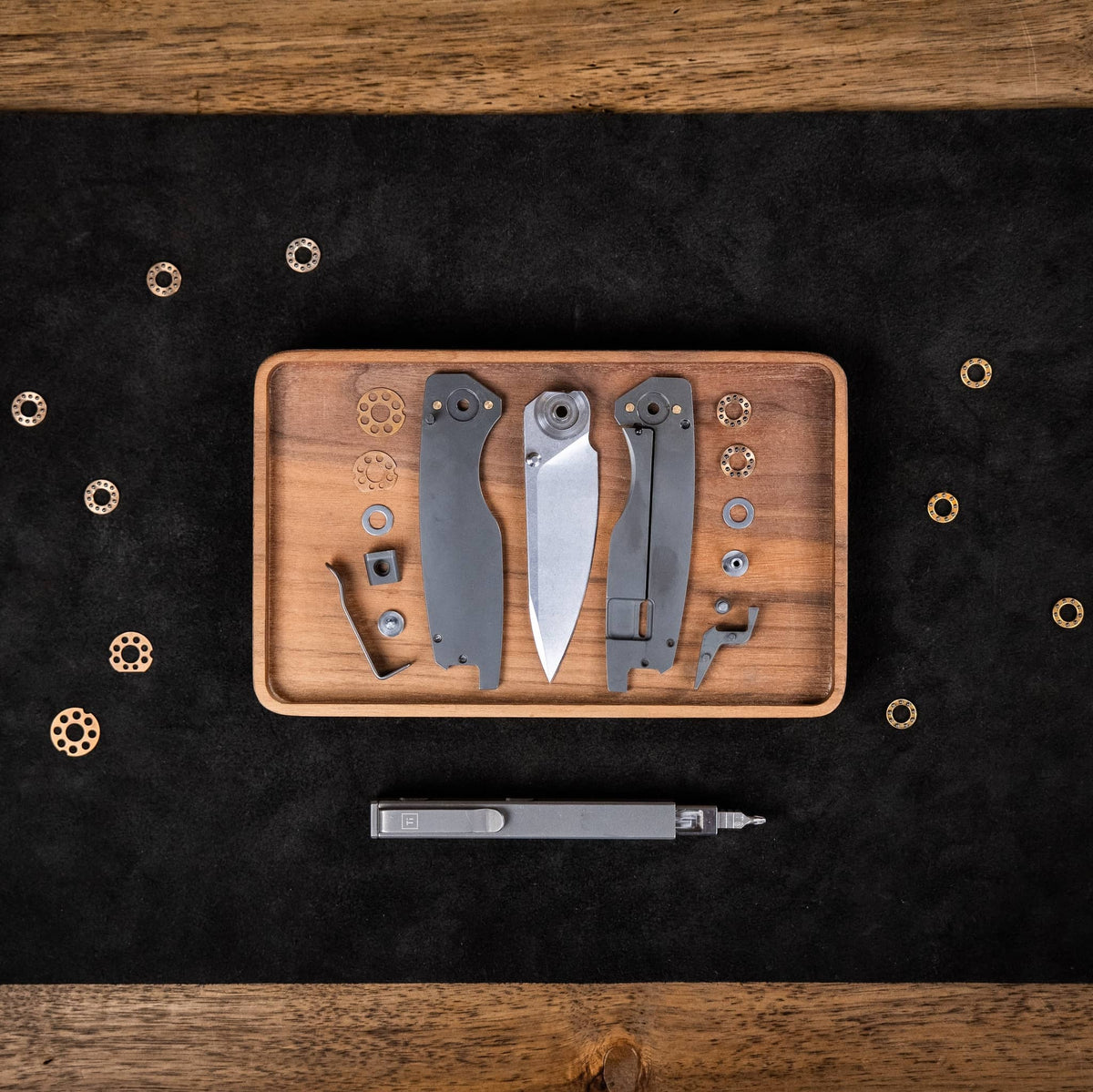 Disassembled Ti Manu knife with titanium handle scales, blade, washers, bearings, and hardware on a wooden tray